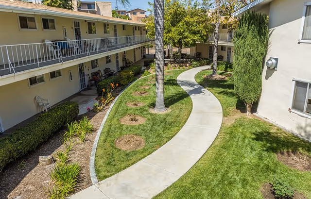 Curved concrete walkway surrounded by green grass, small trees, and shrubs between two beige two-story buildings with balconies and windows under a clear sky.
