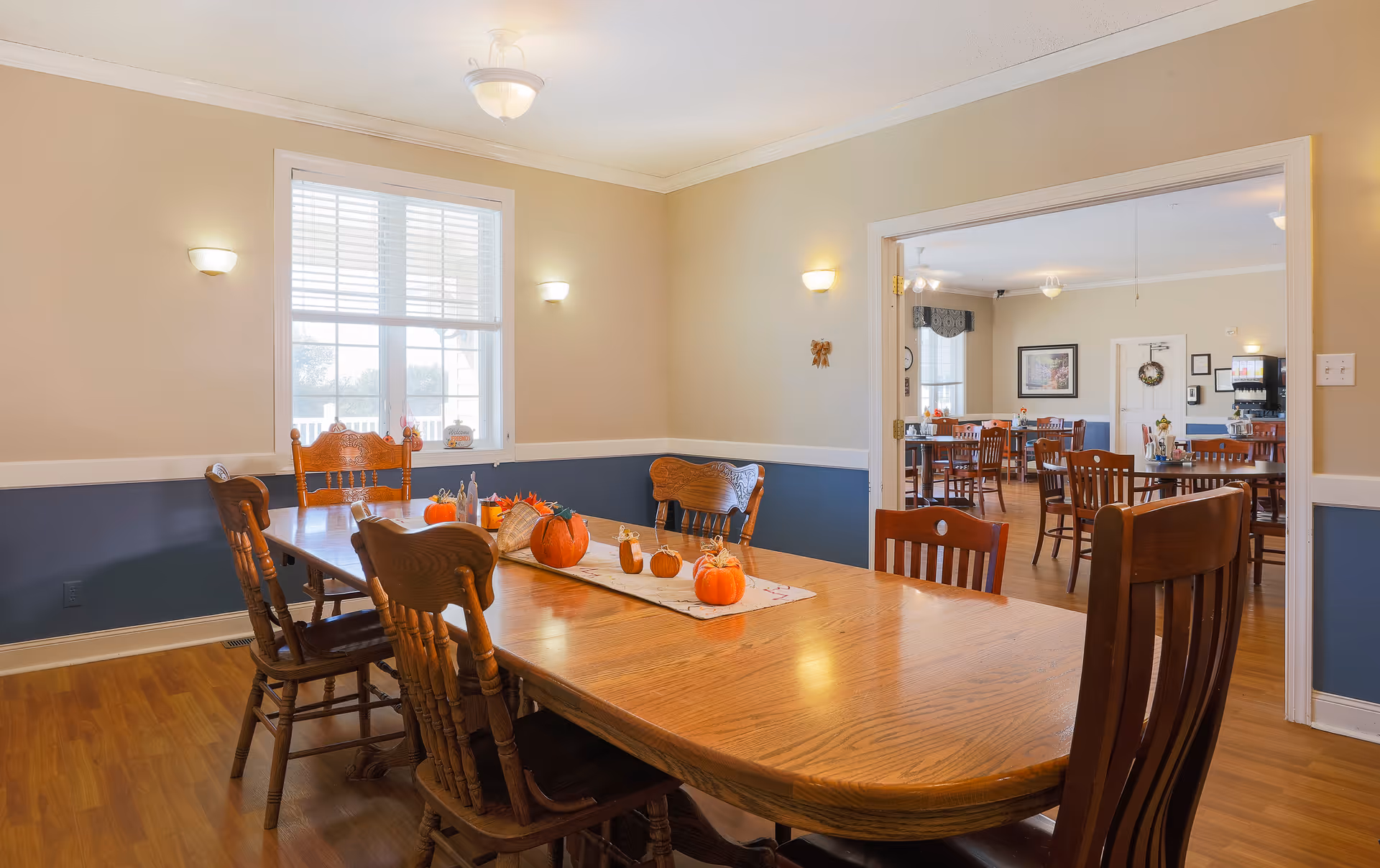 A dining room with a long wooden table surrounded by wooden chairs. The table is decorated with small pumpkins and a table runner. The room has beige walls with a blue wainscot, a window with white blinds, and wall-mounted lights. Through an open doorway, another dining area with more tables and chairs is visible.