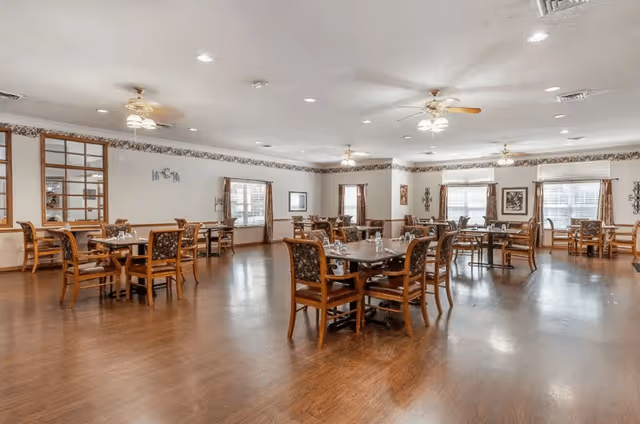 Spacious dining room with multiple wooden tables and chairs arranged neatly on a polished wooden floor. The room has several windows with curtains, ceiling fans with lights, and framed artwork on the walls.