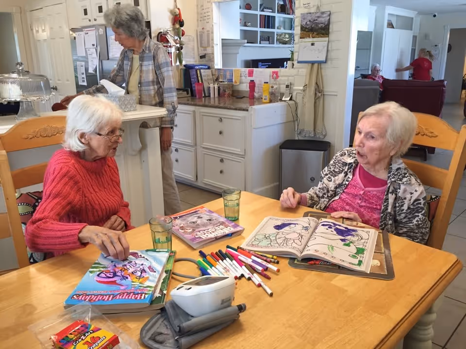 Two elderly women sitting at a wooden table engaged in coloring activities with markers and coloring books. One woman is wearing a red sweater and glasses, while the other is wearing a patterned jacket over a pink top. In the background, another elderly woman stands near a kitchen counter, and a caregiver in red is seen assisting another seated elderly person. The setting appears to be a communal area in a senior living facility with white cabinetry and a cozy atmosphere.