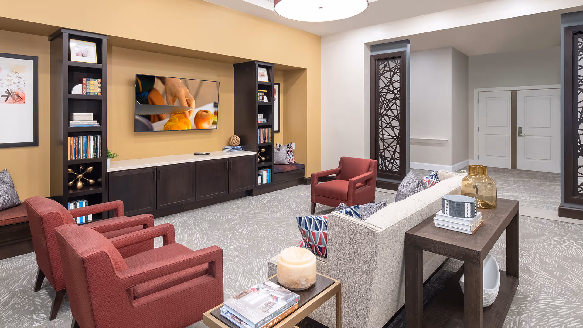 A cozy living room area with a beige sofa adorned with patterned pillows, two red armchairs, and a small table with books and a candle. The room features built-in dark wood shelves and cabinets, a wall-mounted TV showing a close-up of hands cutting fruit, and decorative wall art. The carpet has a light gray pattern, and the walls are painted in warm beige and white tones.