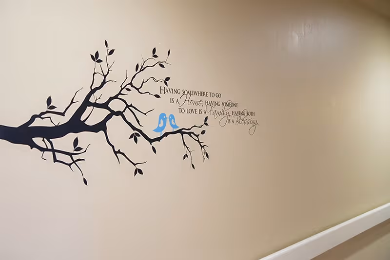 Beige hallway wall decorated with a black tree-branch decal, two blue bird decals and a cursive inspirational quote above a white handrail.