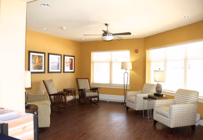 A bright and spacious senior living common area with mustard yellow walls and large windows letting in natural light. The room features two wooden rocking chairs, two upholstered armchairs with a small round table and lamp between them, a floor lamp, and framed artwork on the wall. The floor is dark wood, and a ceiling fan is mounted overhead.