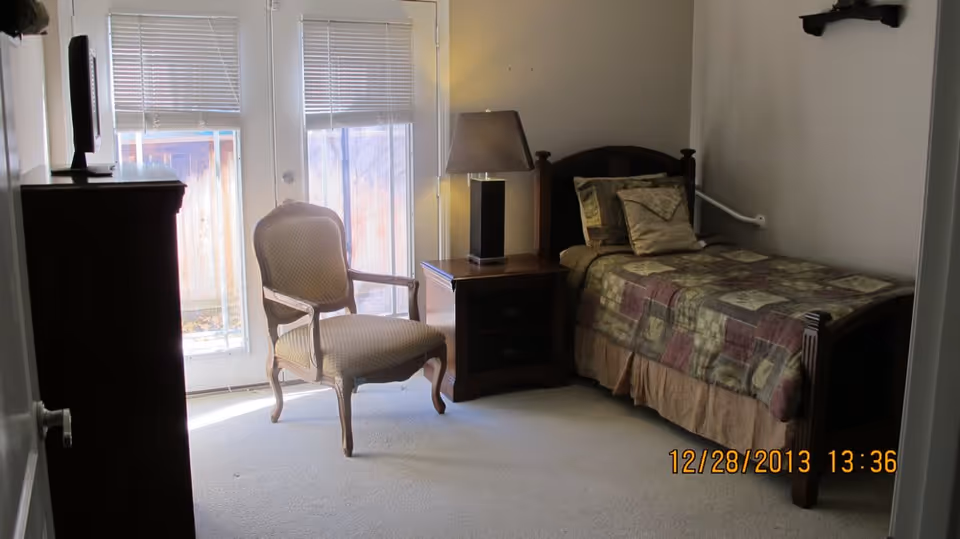 A furnished bedroom in a senior care facility with a bed, chair, dresser, and lamp.