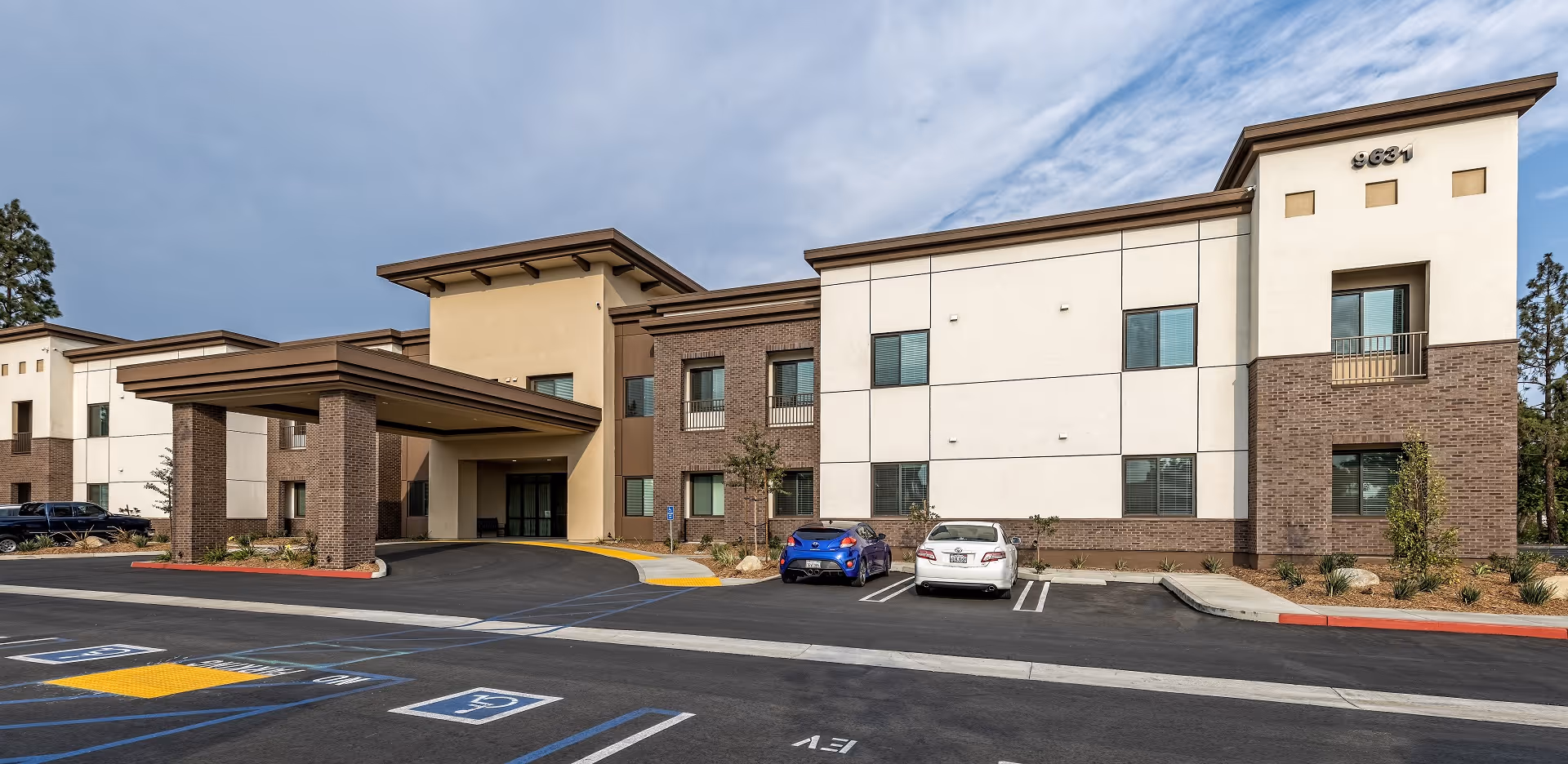 Exterior view of a modern two-story senior living facility building with a covered entrance and several parked cars in front. The building features a combination of light-colored and brick facade with multiple windows and a clear sky above.