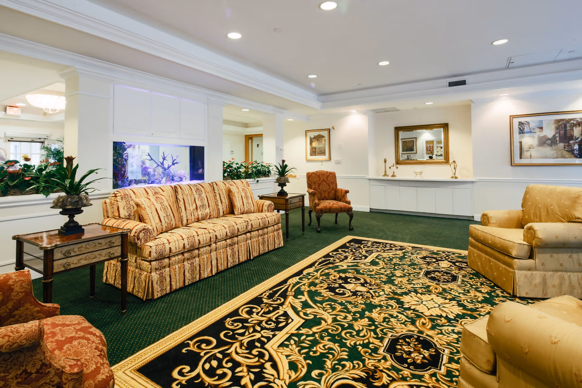 A spacious and elegantly furnished living room area with patterned sofas and armchairs, a large ornate rug with green, black, and gold designs, potted plants on side tables, framed artwork on the walls, a large mirror, and a built-in aquarium with coral and fish.