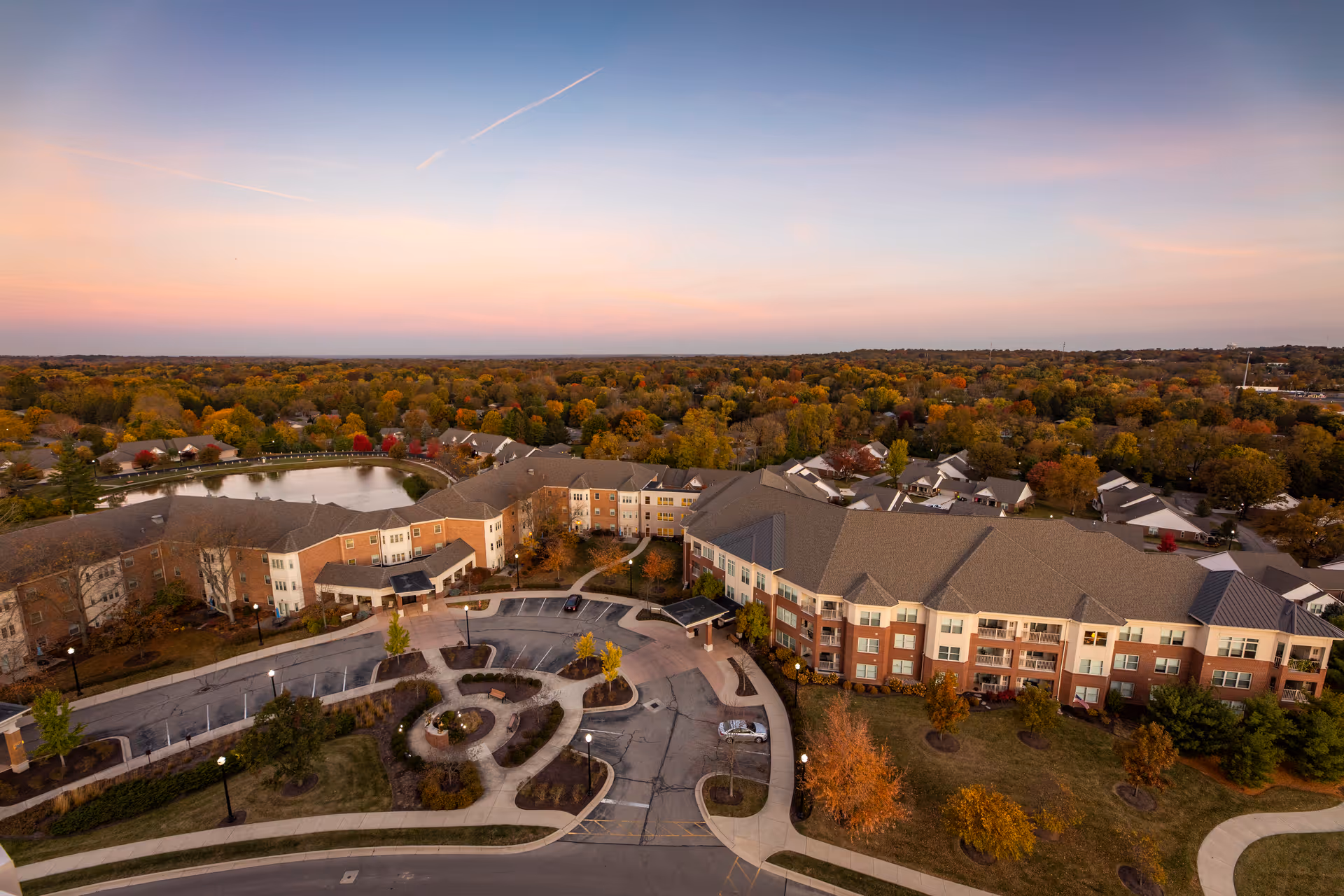 Aerial view of Bethany Village senior living facility during sunset, showing a large multi-story building surrounded by trees with autumn foliage, a pond, parking areas, and curved walkways.