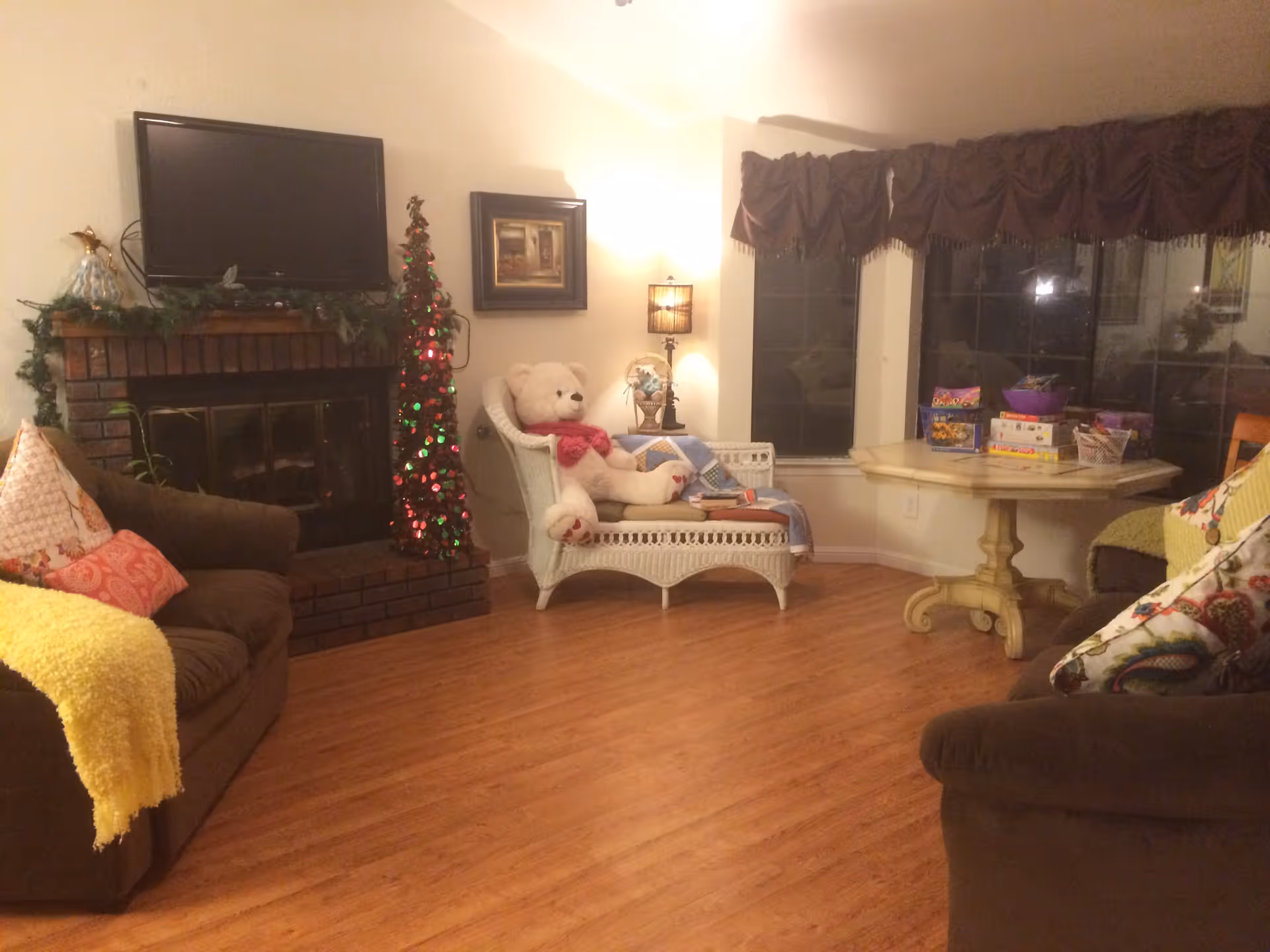 Cozy living room with wooden floor, two brown sofas with colorful pillows and a yellow throw blanket, a white wicker chair with a large teddy bear, a small decorated Christmas tree, a fireplace with a TV mounted above it, a table with board games near large windows with dark curtains, and a lit lamp on a side table.
