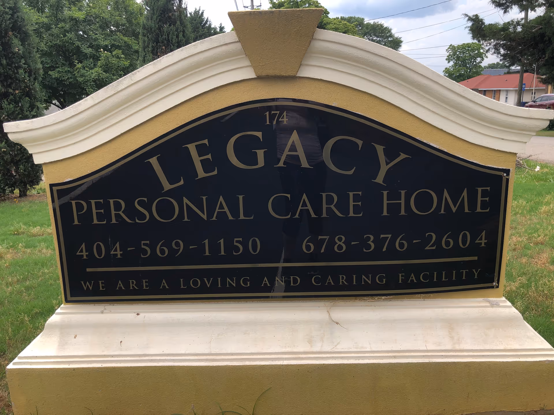 A large outdoor sign for Legacy Personal Care Home with phone numbers 404-569-1150 and 678-376-2604, and the phrase 'We are a loving and caring facility'. The sign is mounted on a yellow and white base with greenery and trees in the background.