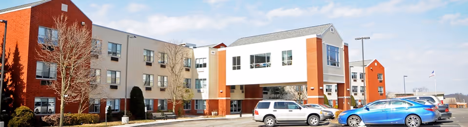 Exterior view of a multi-story senior living facility building with a parking lot in front. Several cars are parked, and there are some trees and shrubs around the building. The sky is partly cloudy.
