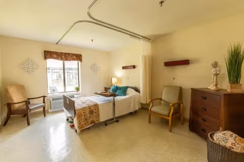 A well-lit healthcare facility room with a single hospital bed featuring white and patterned bedding. The room includes two armchairs, a wooden dresser with decorative items, a window with a valance, and a privacy curtain rail on the ceiling.