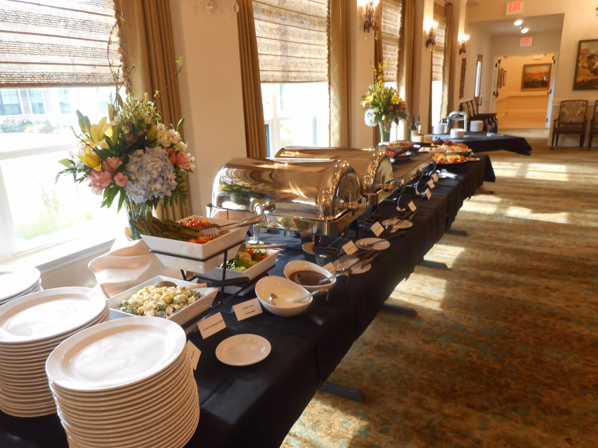 A buffet table set up with chafing dishes, plates, bowls, and serving utensils in a well-lit room with large windows and floral arrangements on the table.