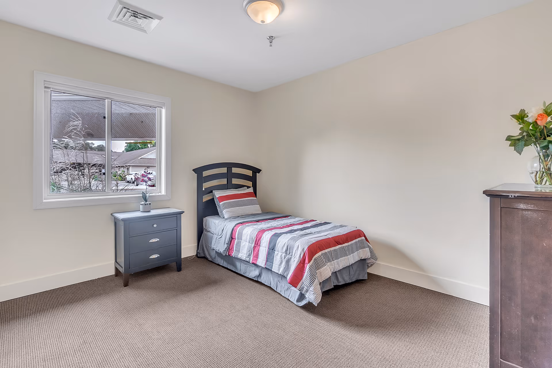 Simple bedroom with a single bed against a beige wall, a small nightstand under a window, and a dresser with flowers.