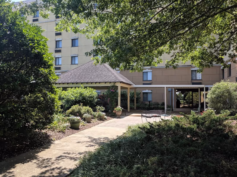 Sunny landscaped courtyard with a covered gazebo, walkway, potted plants and seating in front of a multi-story building.
