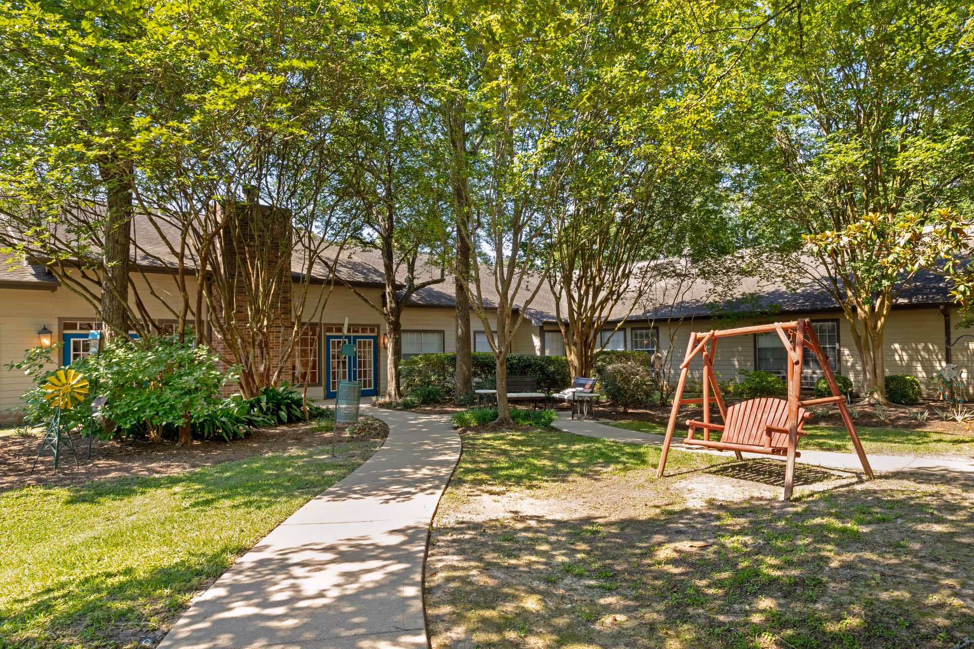 Courtyard with a paved walkway, trees, and a wooden swing in front of a senior living building.
