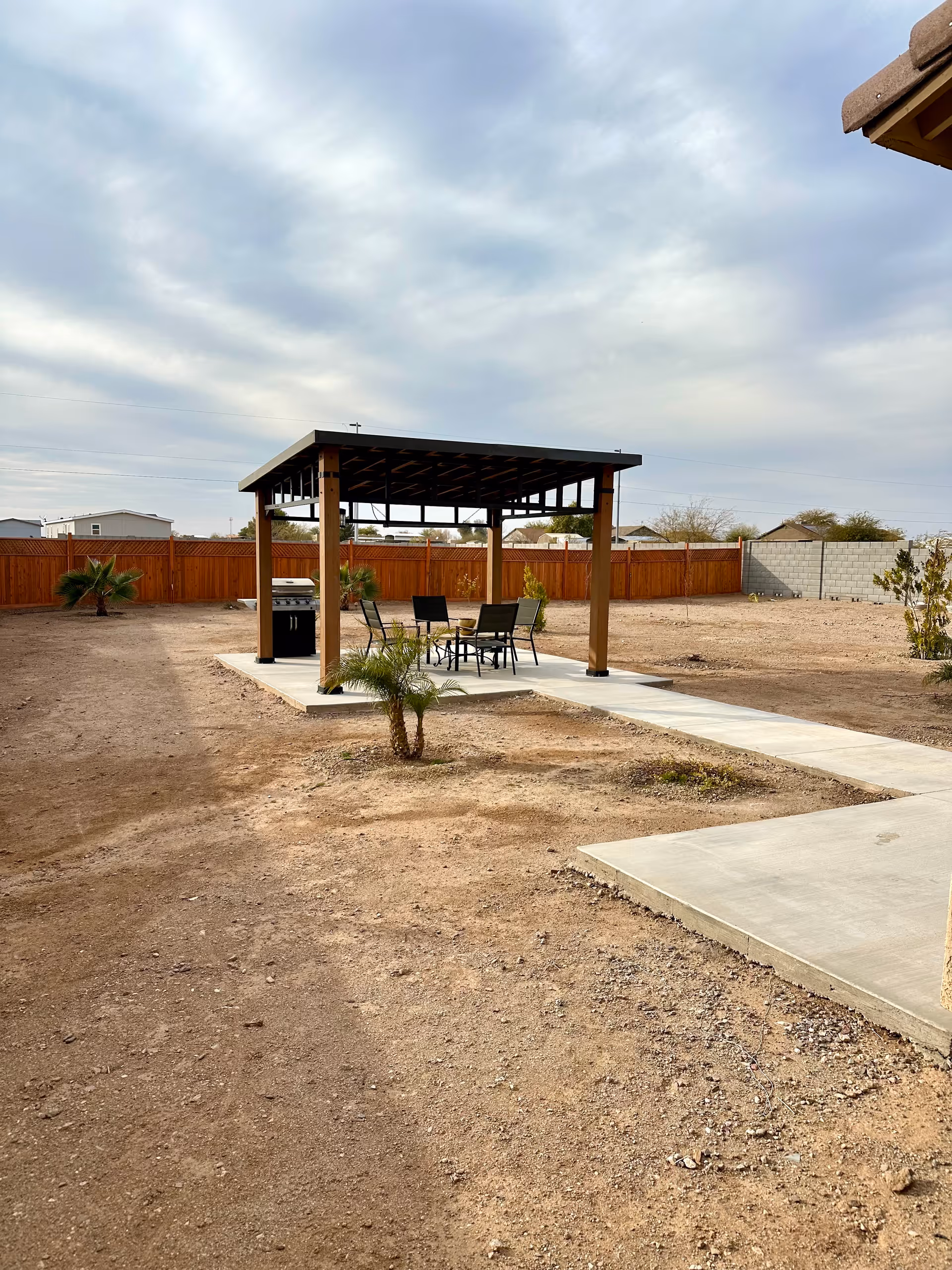A covered pergola with patio furniture and a grill on a concrete pad in a fenced, sandy backyard under a cloudy sky.