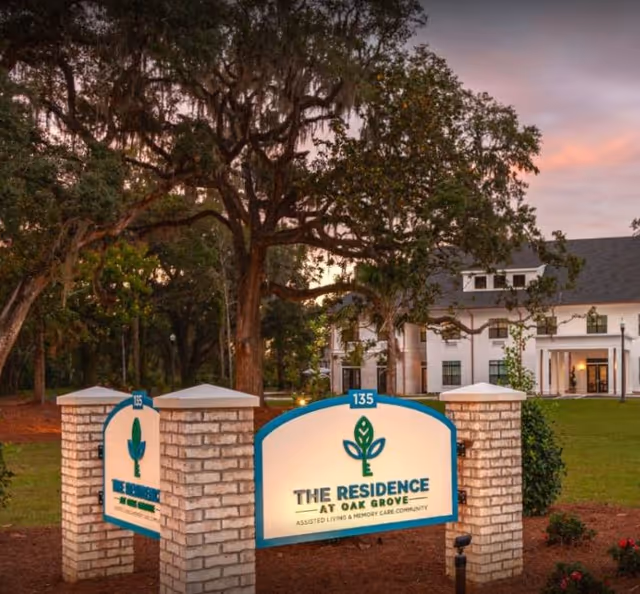 Entrance sign for The Residence at Oak Grove Assisted Living & Memory Care community, mounted on brick pillars with a large tree and a white building in the background during sunset.