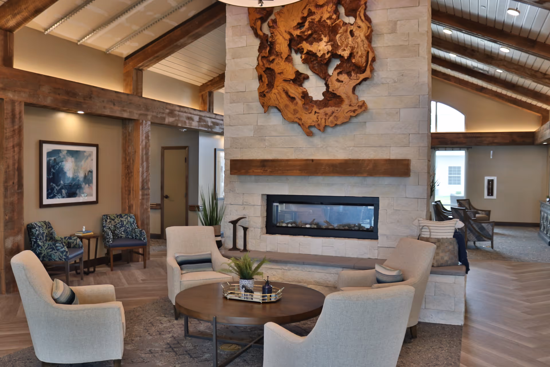 Cozy lounge area with a central stone fireplace, round coffee table and upholstered chairs under exposed wooden beams.