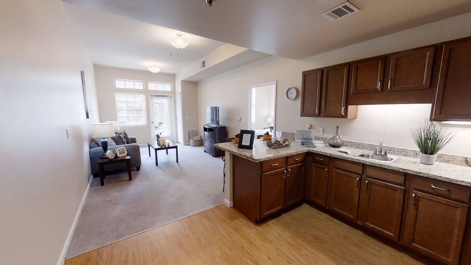 Open-plan living area and kitchen with dark wood cabinets, granite countertops, a sofa and TV by a windowed door.