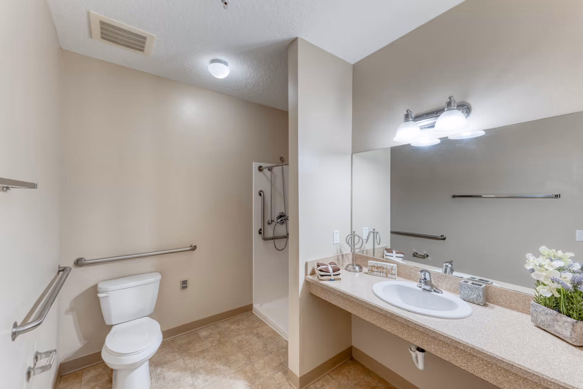 A clean and accessible bathroom with a white toilet, grab bars on the walls, a walk-in shower with a handheld showerhead and grab bars, a large mirror above a countertop with a sink, decorative flowers, and neatly folded towels.