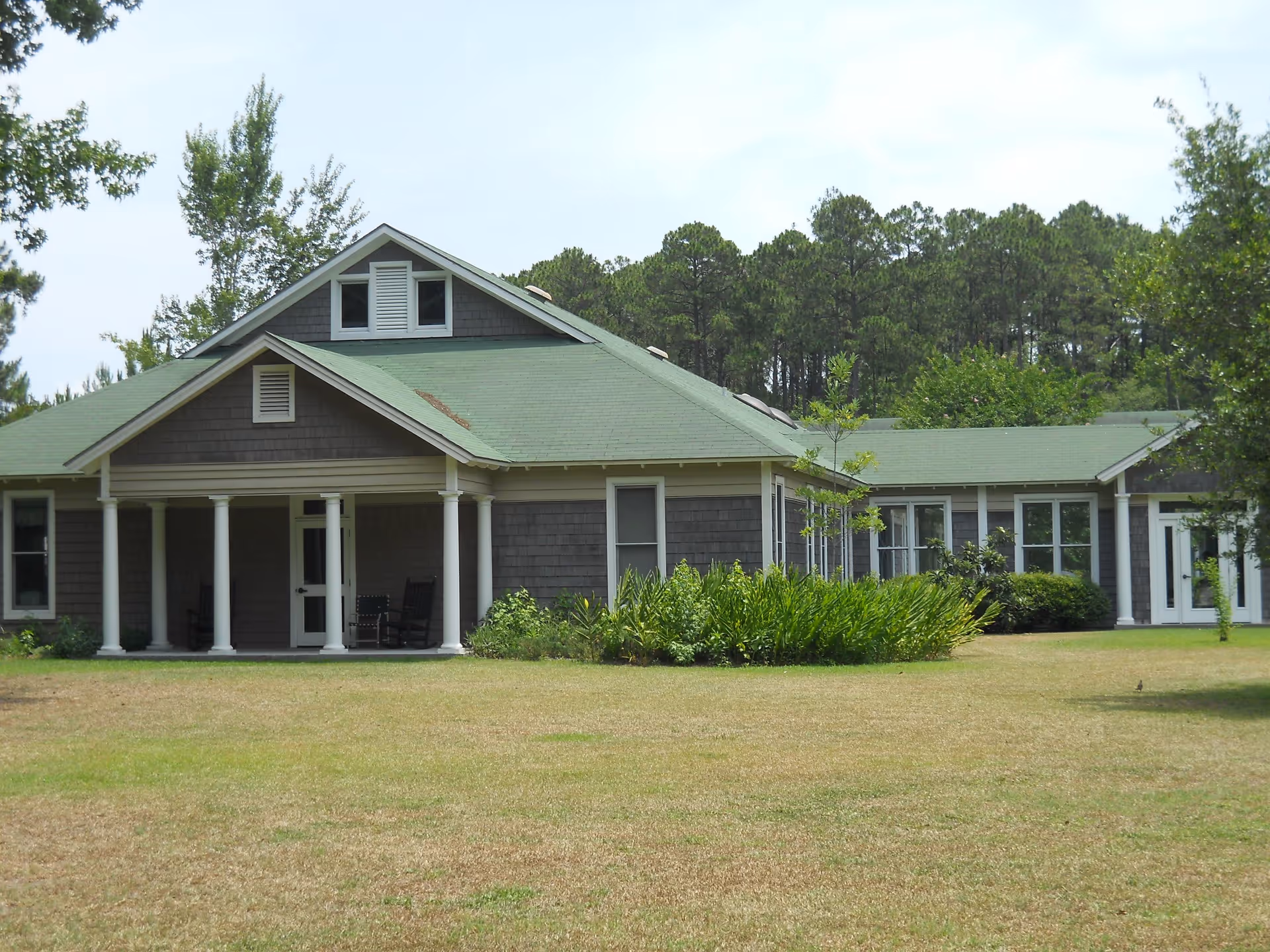 Single-story brown building with a green roof and a columned front porch on a grassy lawn with trees behind.