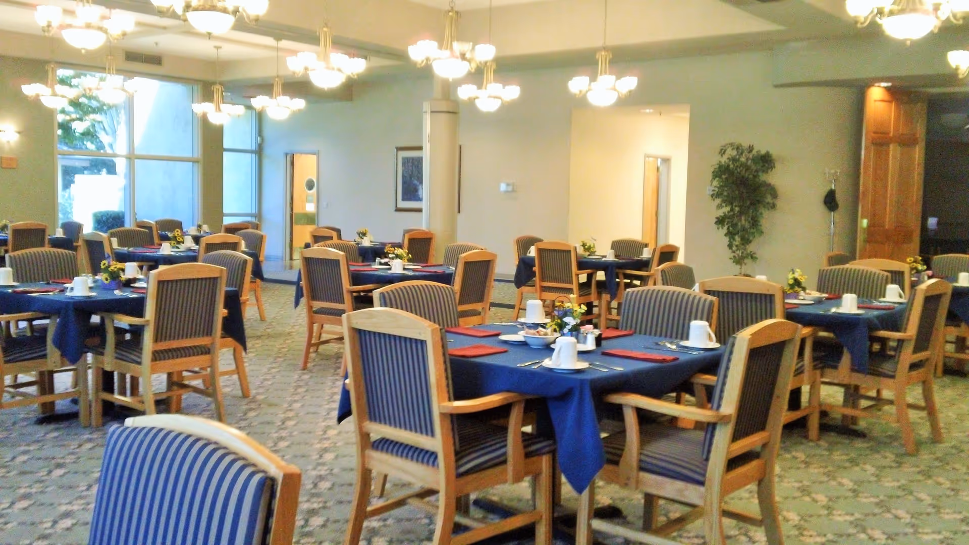 A dining room with multiple round tables covered with blue tablecloths, each set with white cups, plates, silverware, and small flower arrangements. The room has striped wooden chairs, carpeted floor, large windows, and multiple ceiling light fixtures.