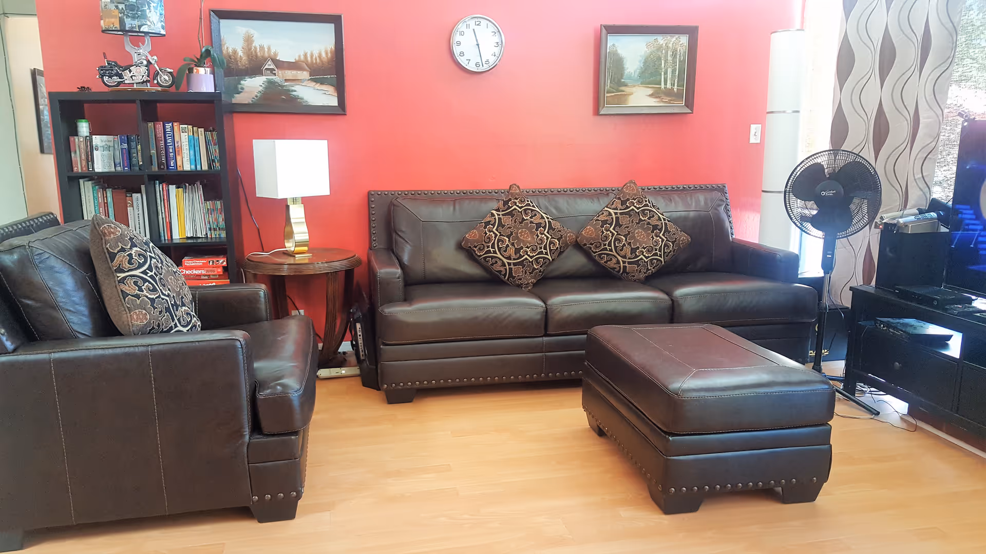 Living room with brown leather sofa, matching armchair and ottoman against a red accent wall with a bookshelf, lamp, and wall clock.