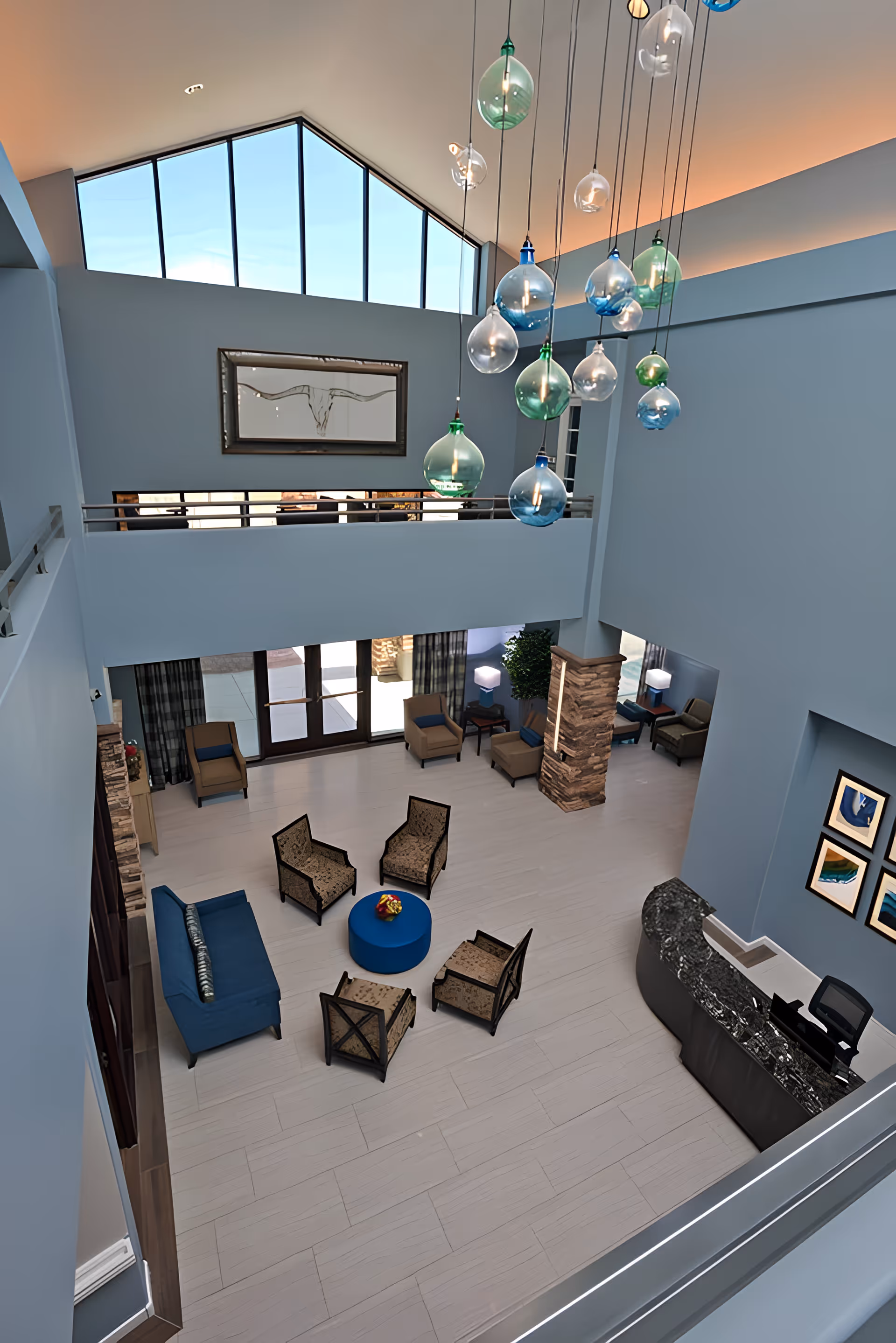 View from above of a spacious assisted living facility lobby with a high ceiling and large triangular window. The room features a modern chandelier with blue and green glass bulbs hanging from the ceiling. Below, there is a seating area with a blue sofa, four patterned armchairs arranged around a round blue ottoman with a flower centerpiece, additional armchairs near the entrance, and a curved reception desk with a computer. The walls are painted light blue and decorated with framed artwork and a large picture of a longhorn skull.