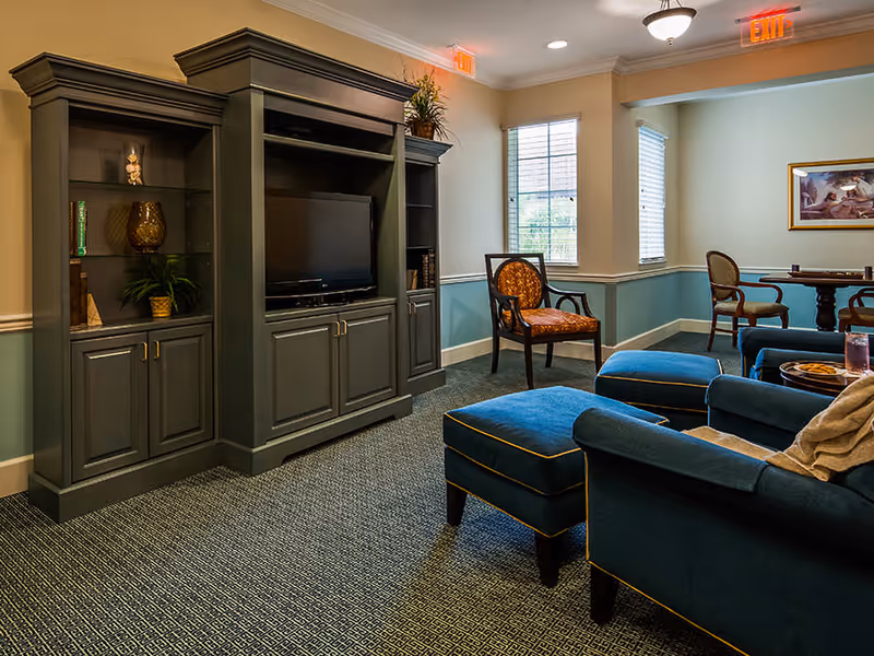A comfortable common living room with upholstered chairs and ottomans facing a TV in a large wooden entertainment cabinet.