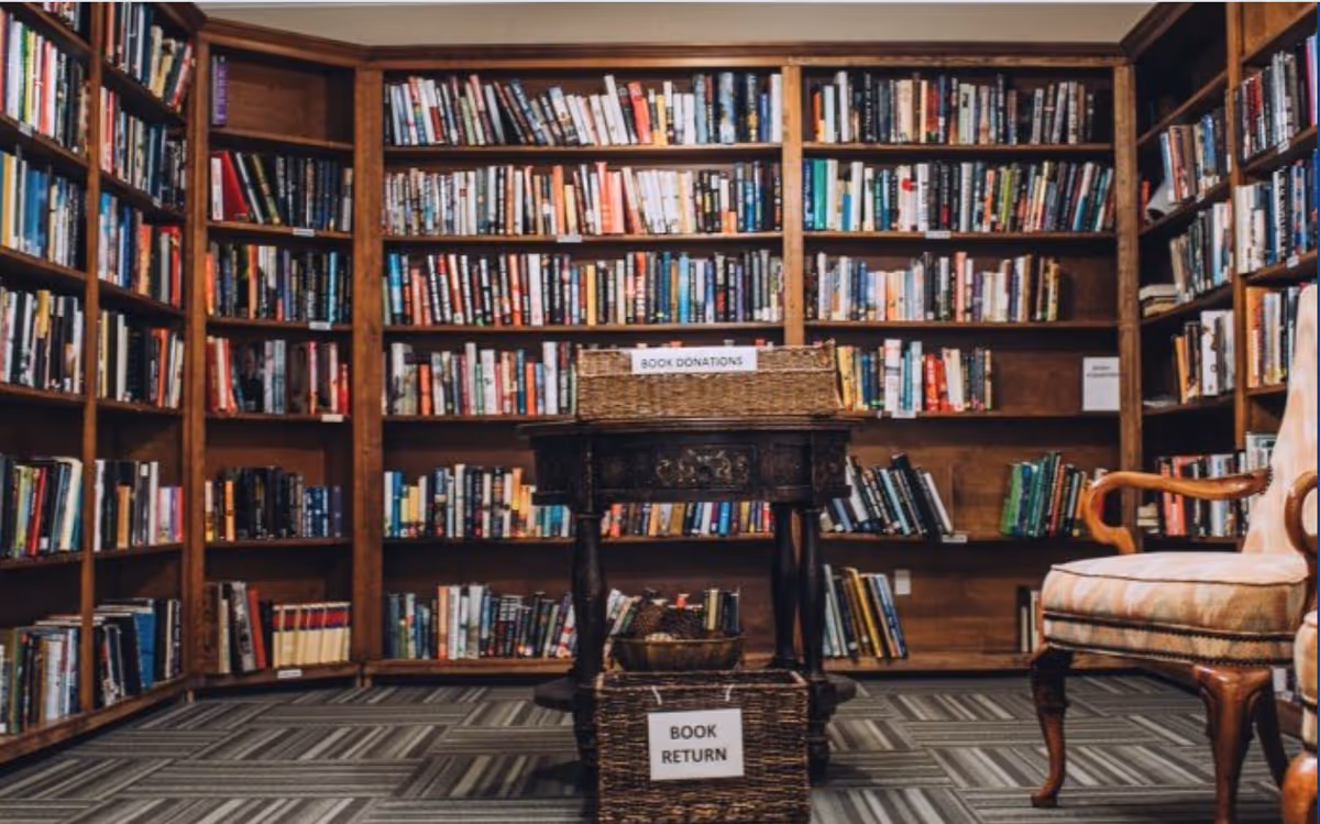 Cozy interior library with wall-to-wall wooden bookshelves, a small table with baskets labeled 'Book Donations' and 'Book Return', and an upholstered chair.