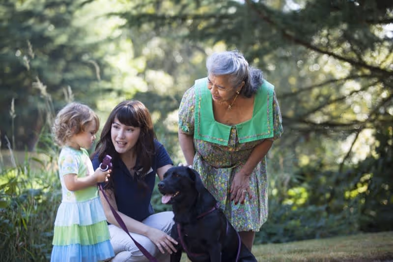 A young woman kneeling on the grass next to a black dog, interacting with a small child in a colorful dress, while an elderly woman in a floral dress looks on, all outdoors in a lush, green park setting.