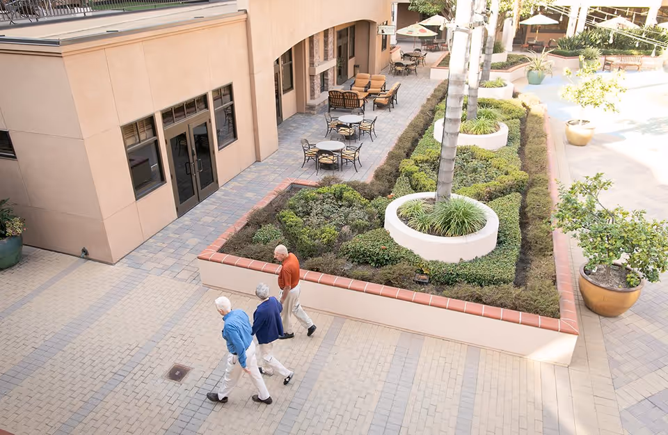 Three elderly individuals walking along a paved outdoor courtyard next to a building with large windows and doors. The courtyard features a raised garden bed with neatly trimmed bushes and palm trees, surrounded by outdoor seating with tables and chairs.