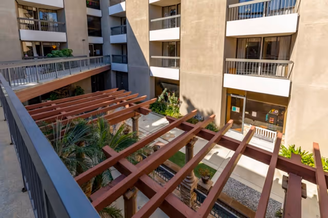 View of an outdoor courtyard area in a multi-story residential building with balconies overlooking a central space featuring wooden pergolas, plants, and a walkway.