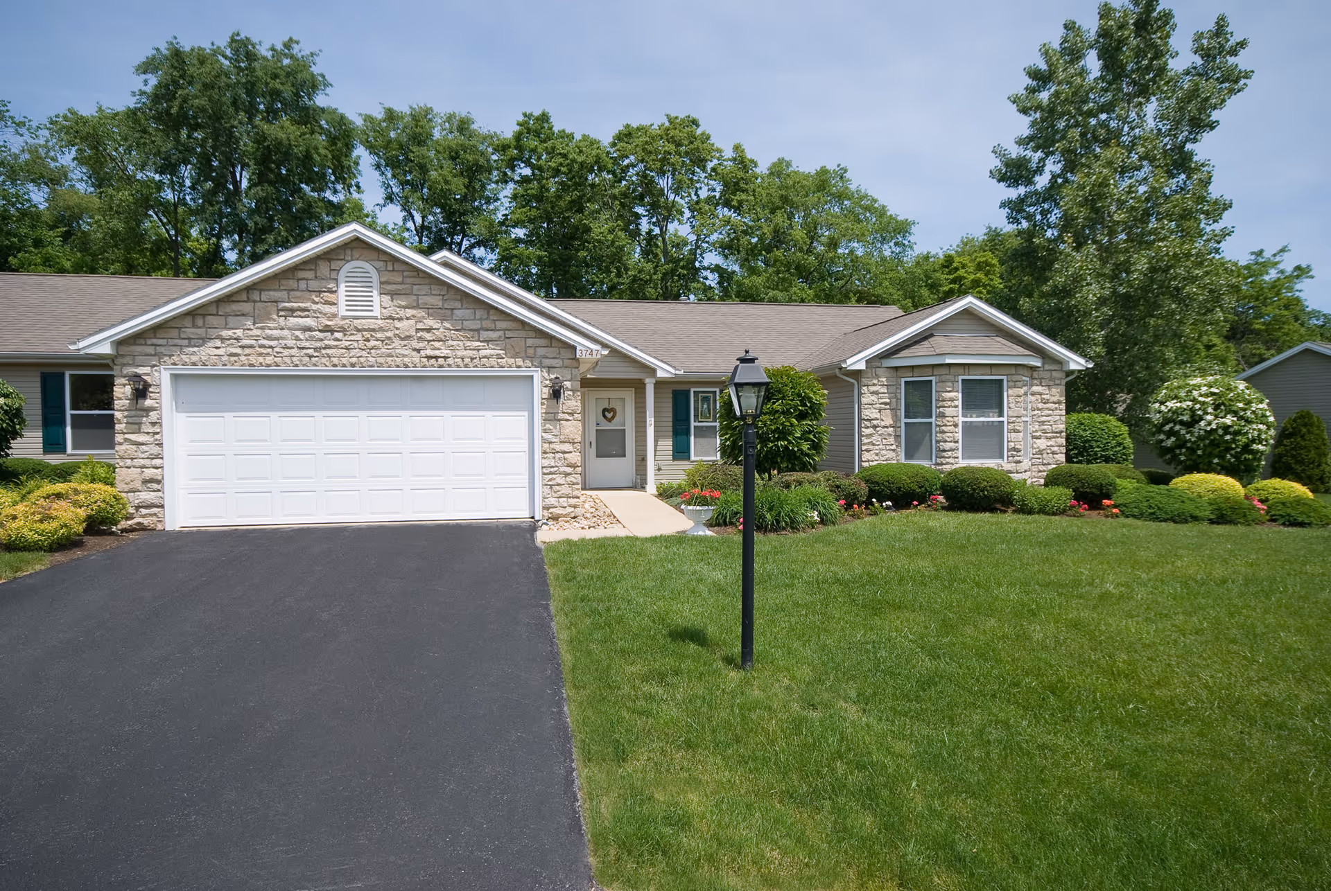 Single-story residential building with a stone facade, a white double garage door, a small front porch with a door featuring a heart-shaped decoration, surrounded by green grass, bushes, and trees under a clear sky.