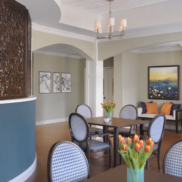 A bright and inviting common area with a square wooden table surrounded by four patterned chairs. A vase with orange tulips is placed on the table. In the background, there is a small seating area with a bench adorned with orange and blue cushions, and a colorful abstract painting on the wall. The room features soft beige walls, decorative wall art, and a modern chandelier hanging from the ceiling.