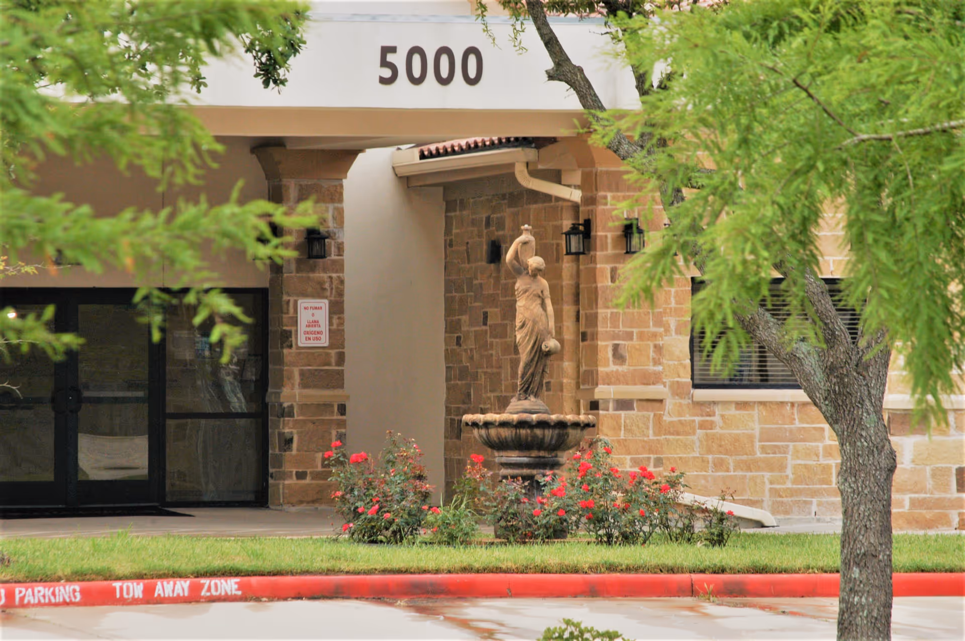 Entrance of a building with the number 5000 displayed above. There is a stone statue fountain surrounded by red flowers and greenery in front of the entrance. The building has a stone facade and there are trees partially framing the view.