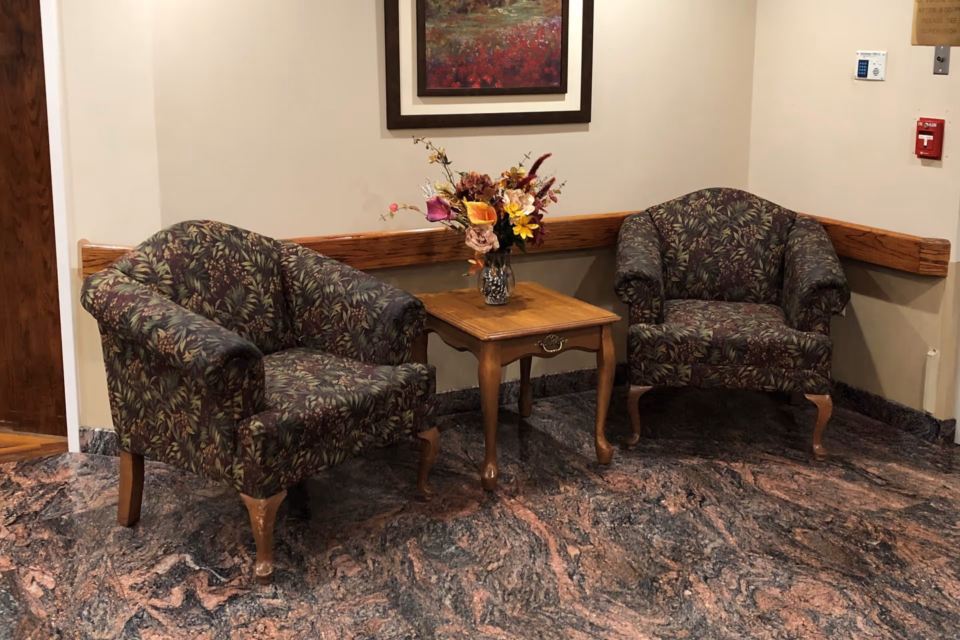 Two patterned armchairs with wooden legs are placed on either side of a small wooden table with a vase of colorful flowers. A framed painting hangs on the beige wall behind the chairs, and there is a fire alarm and a thermostat on the wall to the right.