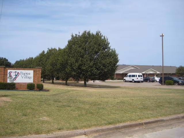 Exterior view of Baptist Village Ada with sign, trees, and parked vehicles.