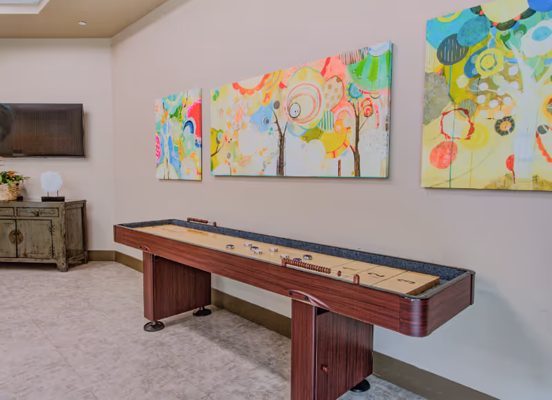 A recreational room featuring a shuffleboard table with pucks on it, colorful abstract paintings on the wall above, a flat-screen TV mounted on the wall, and a rustic wooden cabinet with a plant and decorative item on top.
