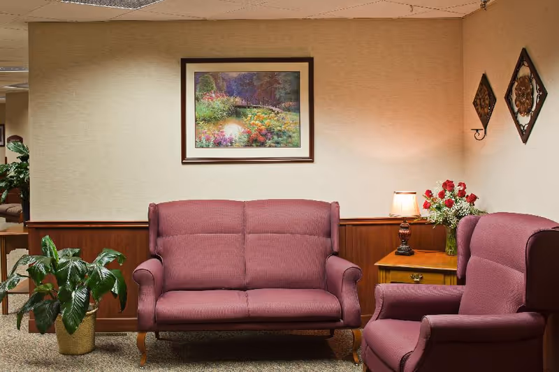 A cozy sitting area with a maroon loveseat and armchair, a wooden side table with a lamp and a vase of red and white flowers, a green potted plant on the floor, and a framed painting of a colorful garden on the beige wall.