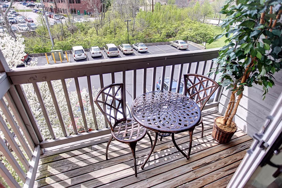 A small wooden balcony with a round metal table and two matching chairs. There is a potted plant with green leaves on the right side. The balcony overlooks a parking lot with several cars and some greenery in the background.