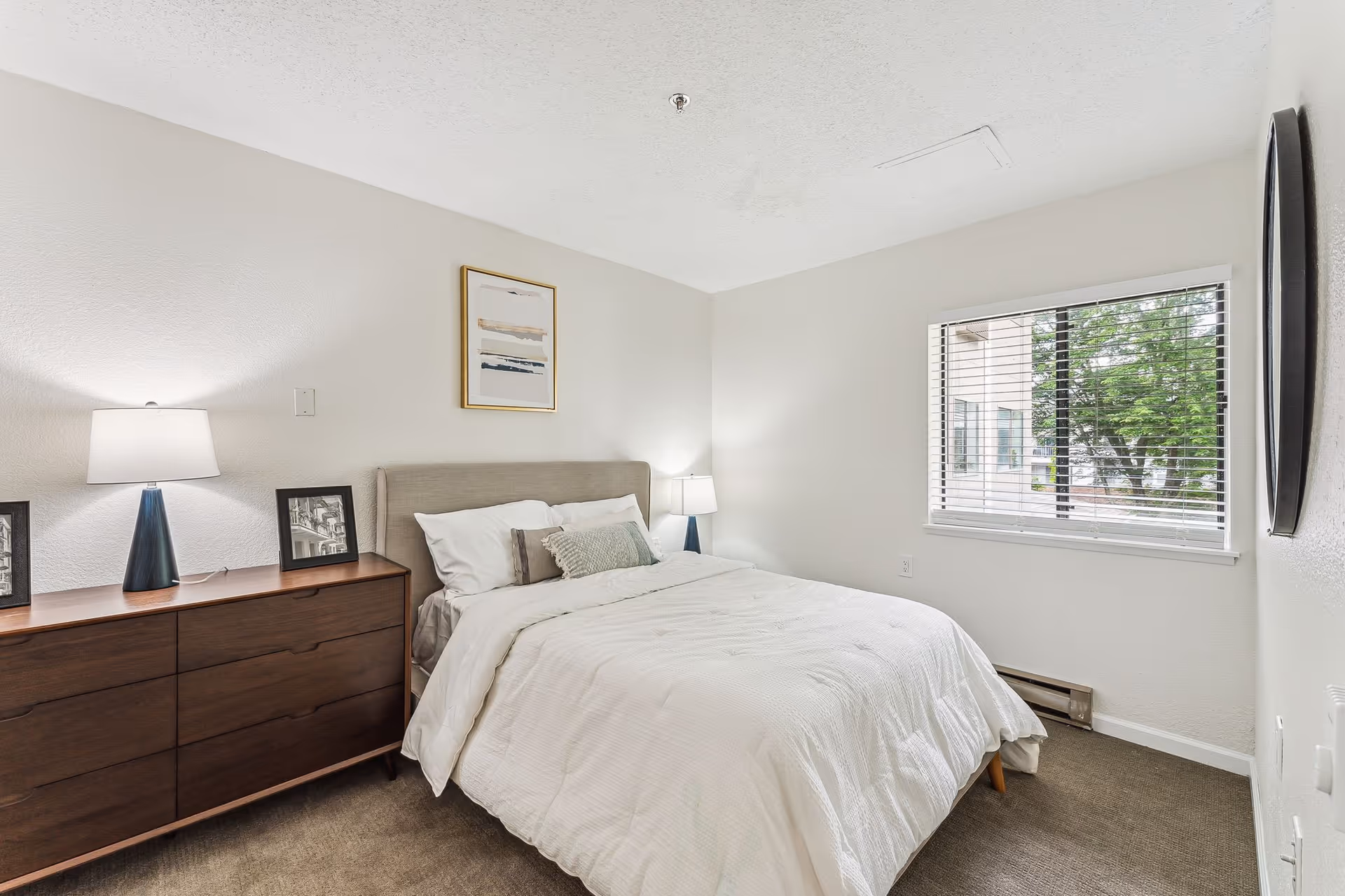 A bright bedroom with a neatly made bed featuring white bedding and multiple pillows. There is a wooden dresser with two table lamps and framed pictures on it. A window with blinds lets in natural light, and a framed abstract artwork hangs above the bed.