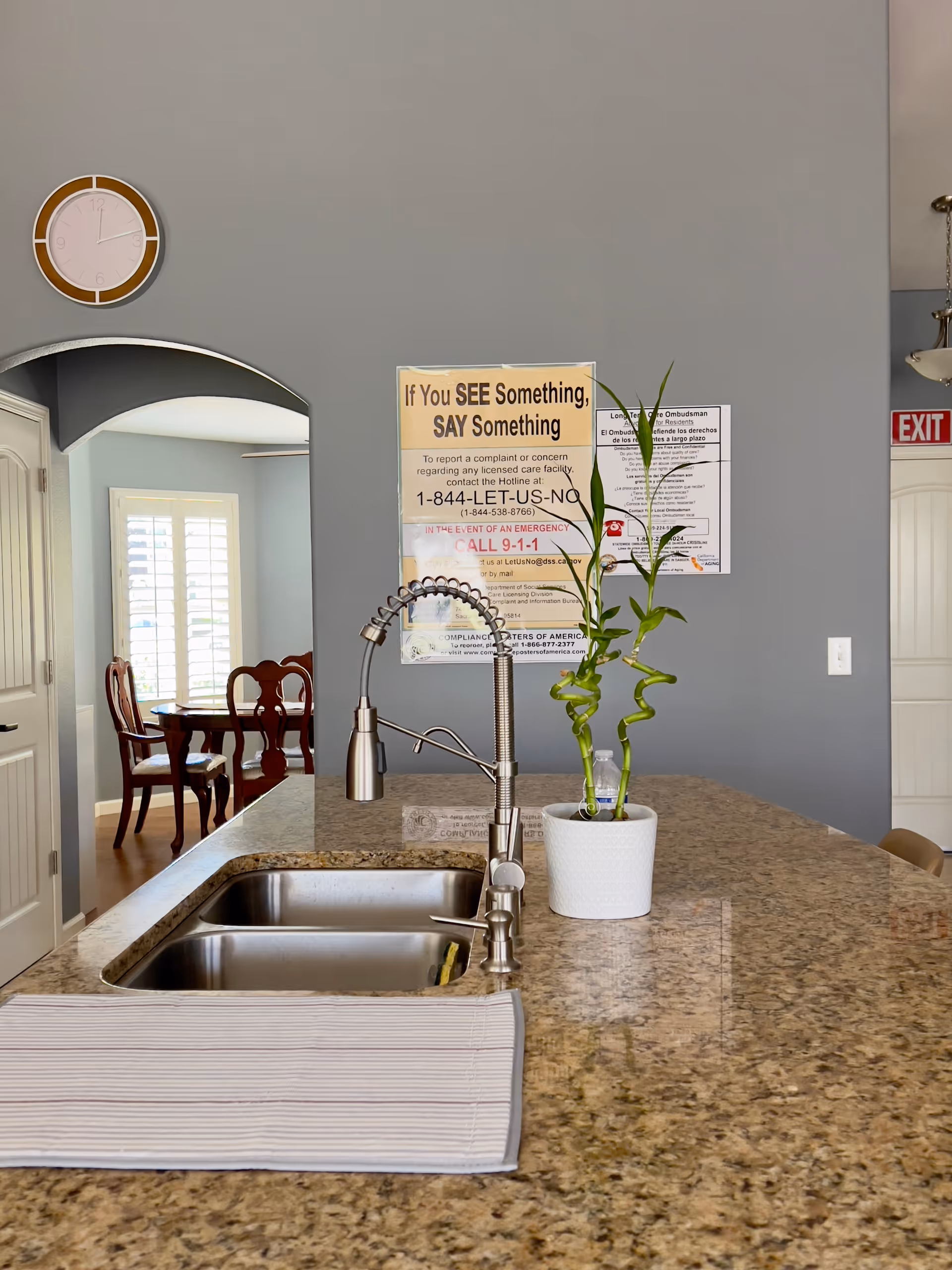 Interior view of a kitchen area featuring a granite countertop with a double stainless steel sink and a modern flexible faucet. A white potted plant with green bamboo stalks is placed on the countertop. In the background, there is a gray wall with informational posters, a round wall clock, and an arched doorway leading to a dining area with wooden chairs and a table. An exit sign is visible on the right side.