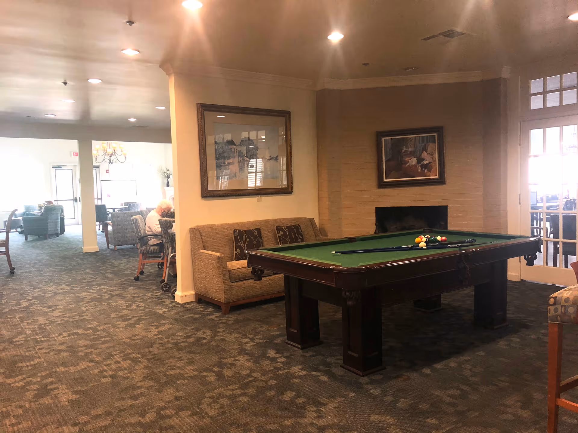 An interior common area in a retirement community featuring a pool table with cues and balls set up, a beige couch with patterned pillows, framed artwork on the walls, and additional seating areas in the background with elderly residents sitting and conversing.