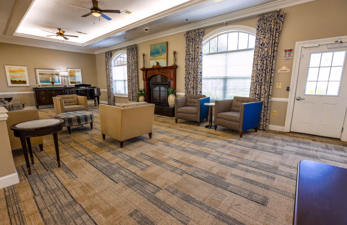 A spacious and well-lit living room area with beige and blue armchairs, a patterned carpet, a wooden fireplace with decorative items, large windows with patterned curtains, ceiling fans, and framed artwork on the walls. There is also a piano and a sideboard in the background.