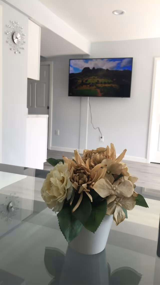 A modern living room with a wall-mounted flat screen TV displaying a landscape image. In the foreground, there is a white pot with artificial flowers on a glass table. The room has light-colored walls and flooring, a decorative wall clock, and a doorway leading to another room.