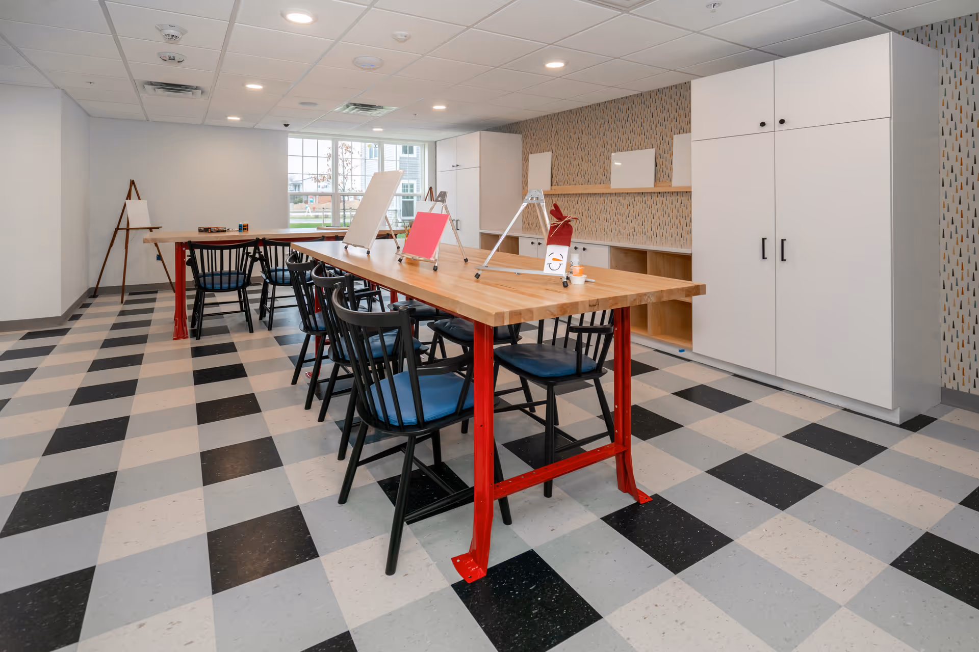 A bright, spacious activity room with a checkered black, white, and gray floor. There are two long wooden tables with red metal legs, surrounded by black chairs with blue cushions. On the tables are small easels holding blank canvases and a few art supplies. The back wall features white cabinets and a patterned wallpaper. Large windows allow natural light to fill the room.