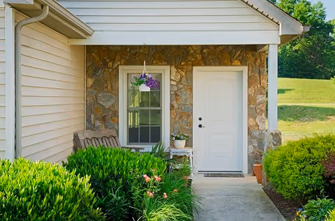 Front entrance of a house with a white door and a window beside it. The exterior wall is made of stone and siding. There is a small porch area with a hanging purple flower pot, a bench, and a small table with a potted plant. Green bushes and plants surround the walkway leading to the door.