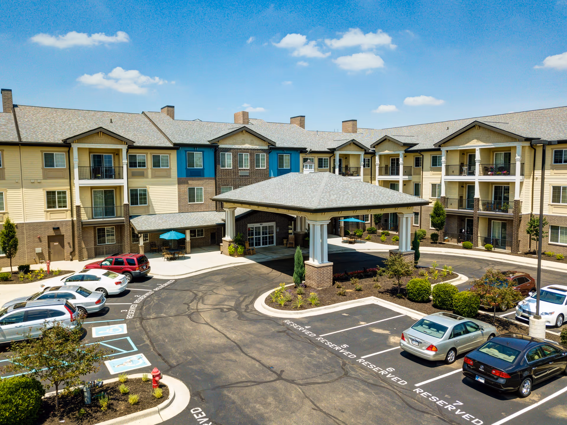 Exterior view of Independence Village of Carmel, showing a three-story residential building with balconies, a covered entrance area, and a parking lot with several cars parked. The sky is clear with a few clouds.