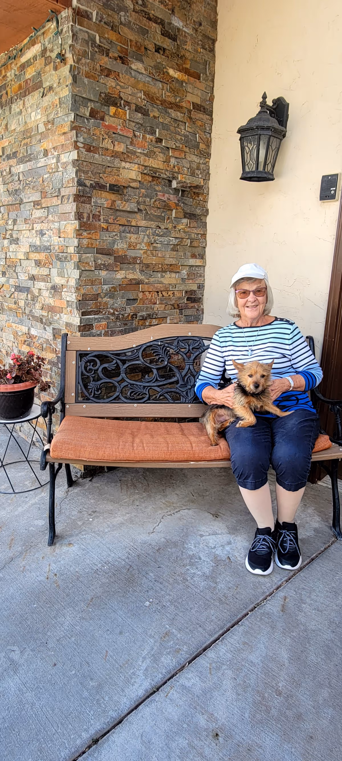 An elderly woman wearing a white cap, glasses, and a striped long-sleeve shirt sits on a cushioned bench outside a building. She is holding a small brown dog on her lap. The bench is positioned against a wall with a stone corner and a black outdoor lantern above. There is a small table with a potted plant next to the bench.